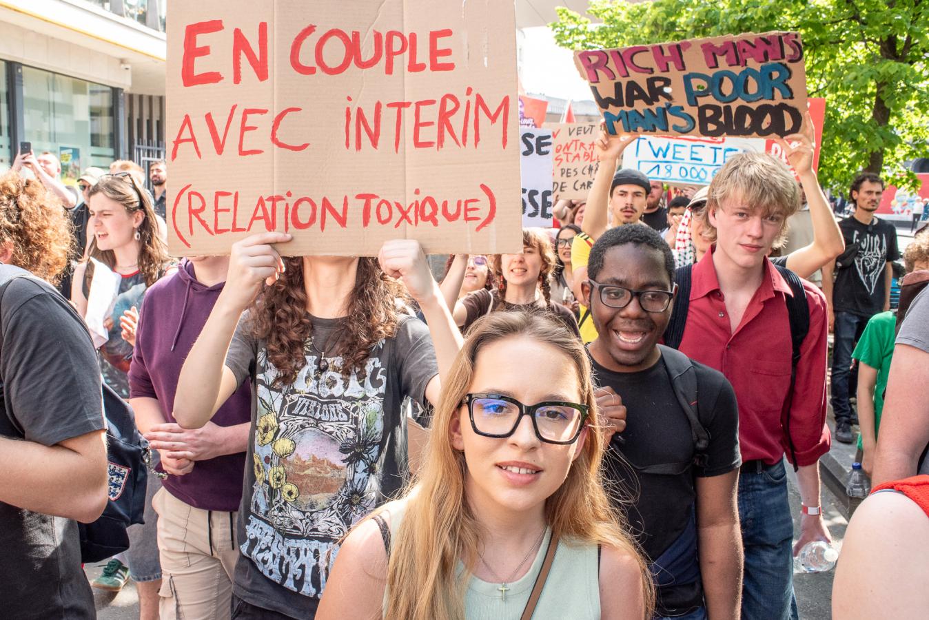 Un groupe de jeune lors de la marche. Pancartes : "En couple avec interim (relation toxique)" et "Rich man's war, poor man's blood"