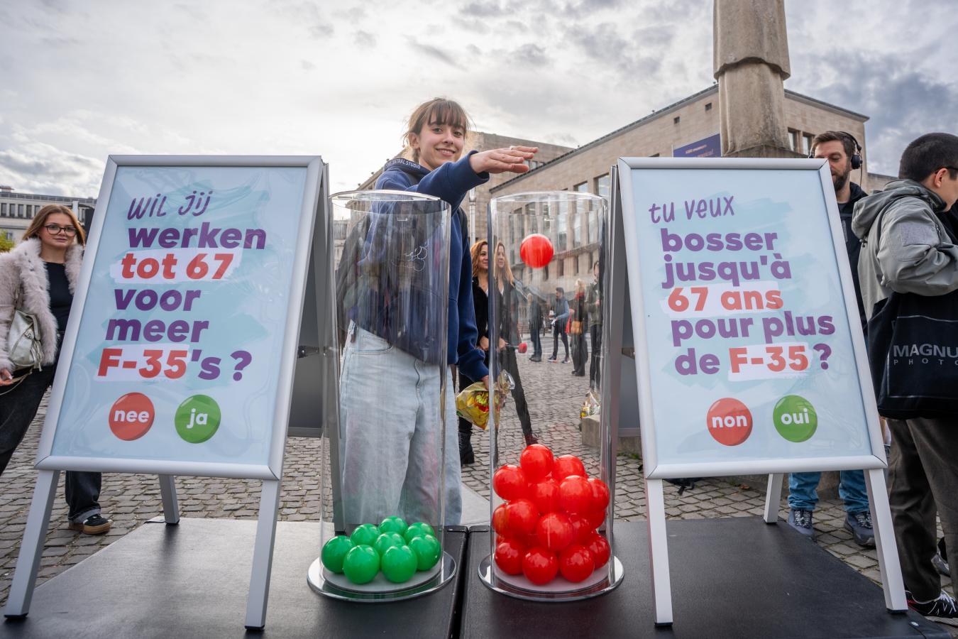 Une jeune femme place une boule rouge dans le bac "non" pour voter sur la question "Tu veux bosser jusqu'à 67 ans pour plus de F-35 ?"