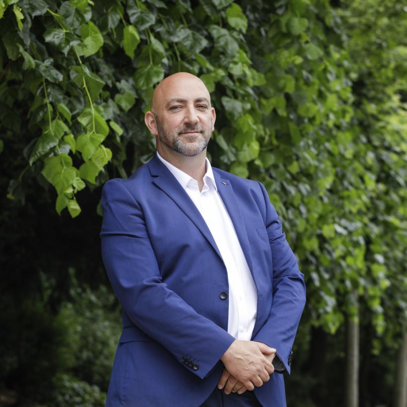 Gaby Colebunders se tient devant une rangée d'arbres. Il porte un blazer bleu foncé et une chemise blanche. Il tient ses mains devant son ventre et sourit avec précaution. 