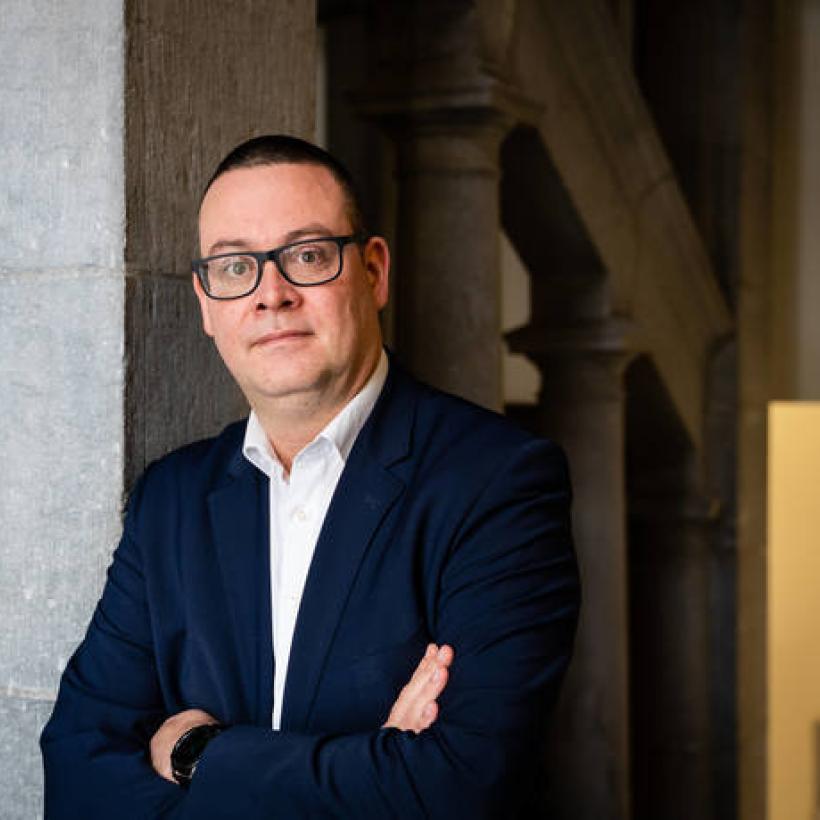 Raoul Hedebouw porte une veste sombre et une chemise blanche. Il est appuyé contre un mur, les bras croisés.