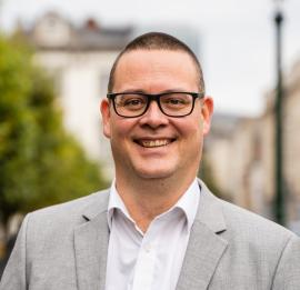 Raoul Hedebouw marche dans la rue à côté du parc Warren. Raoul sourit et regarde la caméra. Il porte un blazer gris et une chemise blanche.