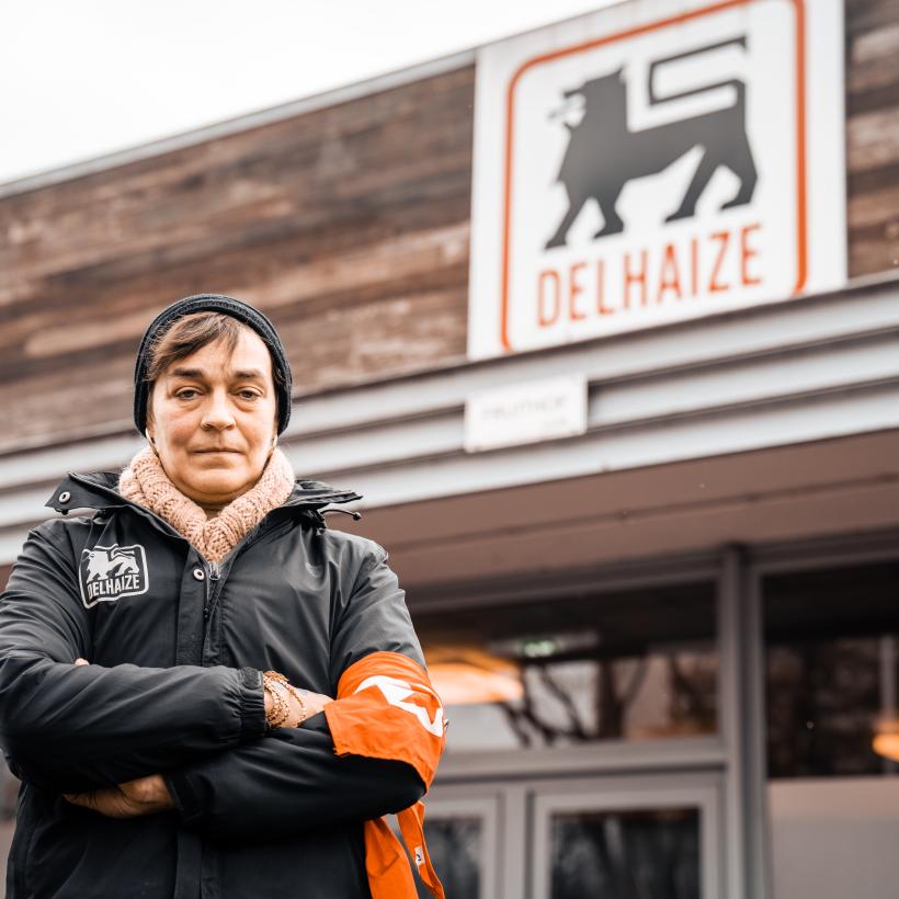 Une travailleuse se tient devant un supermarché surplombé d'un grand panneau « Delhaize ». Elle est habillée chaudement. Elle a les bras croisés et le regard déterminé.