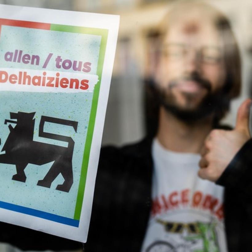 Un jeune homme placadre une affiche du PTB « Tous Delhaiziens » derrière une vitre. Il fait un pouce en l'air.