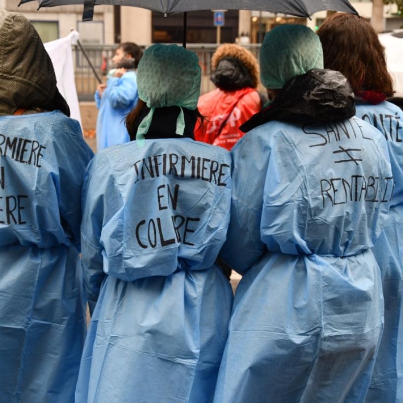Des travailleuses des soins lors d'une manifestation du secteur des soins. Elles sont de dos et sur leurs blouses, on peut lire : "Infirmière en colère. Santé n'est pas égal à rentabilité"
