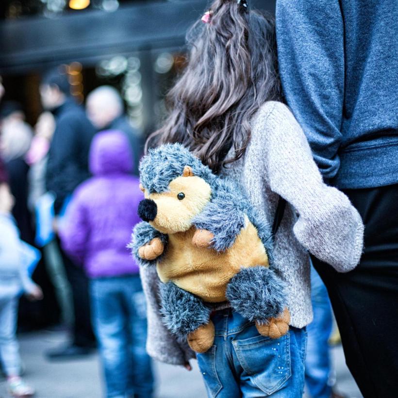 Une petite fille de dos portant un sac à dos en forme de hérisson et tenant la main d'un adulte